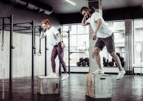 Athletic man and woman practicing lunges together with dumbbell on box in modern gym