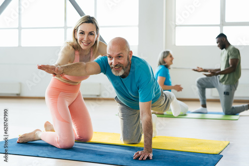 Woman helps mature man to stand in bird dog pose in gym