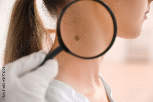 Dermatologist examining mole on young woman's neck with magnifier in clinic, closeup