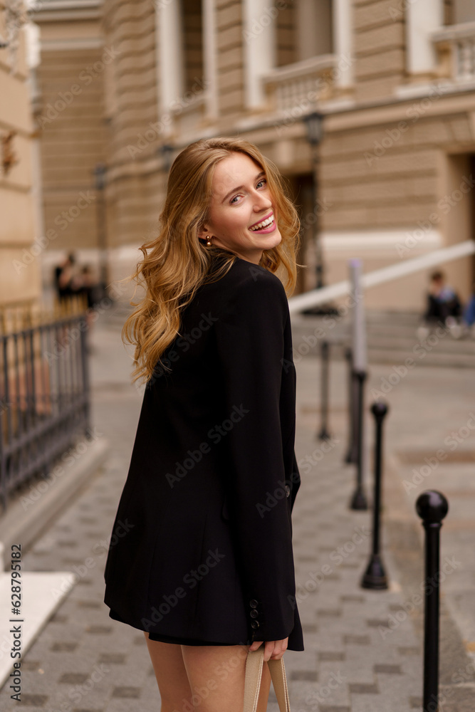 Beautiful smiling woman in elegant black suit  posing outdoor in old european city.  Blond wavy hairs, perfet skin.