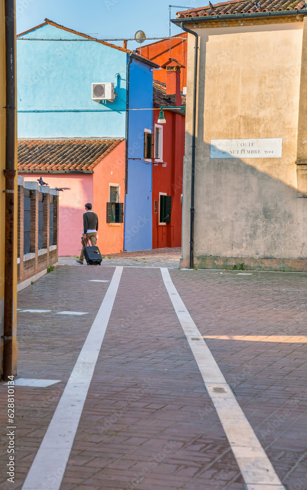 Unidentifiable rear view of a solo traveler with a trolley bag on the narrow colorful alleys of Burano, Venice