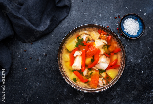 Fish soup with cod, red paprika, potatoes, tomatoes and parsley in soup bowl on black table background, top view