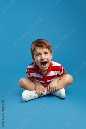 Cute boy wearing red striped shirt, screaming loudly while sitting with crossed legs on studio floor