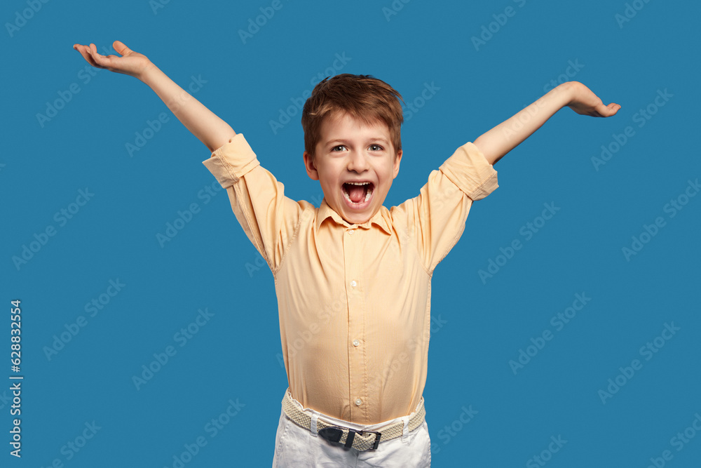Funny little kid boy in beige shirt, posing with raised arms isolated over blue background Funny little kid boy in beige shirt, posing with raised arms isolated over blue background