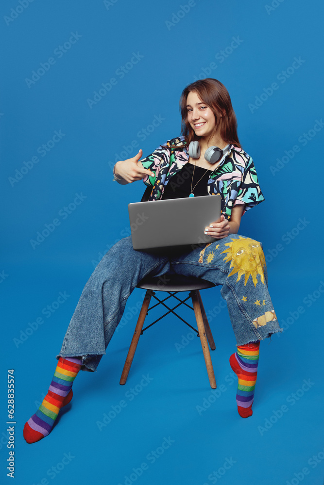 Fototapeta premium Positive lady in trendy shirt and blue jeans pointing index finger on laptop while sitting on chair, isolated over blue background. Student wearing colorful socks and having fun in studio