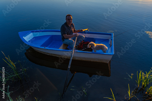 A 60-year-old Caucasian man on a blue wooden boat with oars in his hands with a poodle dog lit by the sun at sunset.