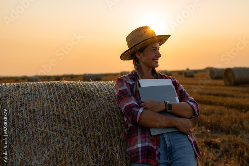 Smiling farmer woman with tablet, folders, and straw hat posing in her hay field amidst rolled bales. Sunsets and fields provide a picturesque backdrop © DusanJelicic