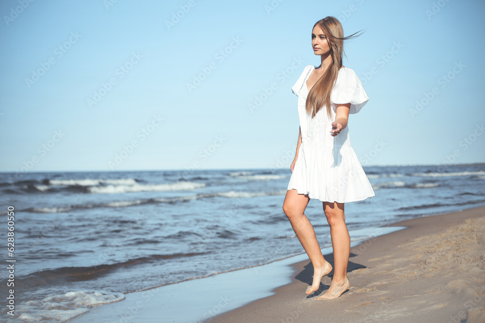 Happy smiling beautiful woman is walking on the ocean beach in a white summer dress