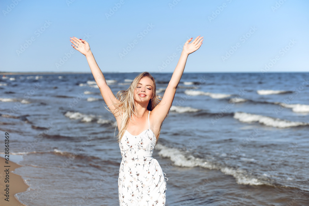 Happy blonde beautiful woman on the ocean beach standing in a white summer dress, raising hands Happy blonde beautiful woman on the ocean beach standing in a white summer dress, raising hands