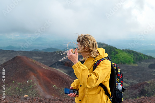 Hiking on tallest volcano in Continental Europe - Etna. Young smiling woman in yellow raincoat in the mountain peak of panoramic view of Mount Etna in a windy day.