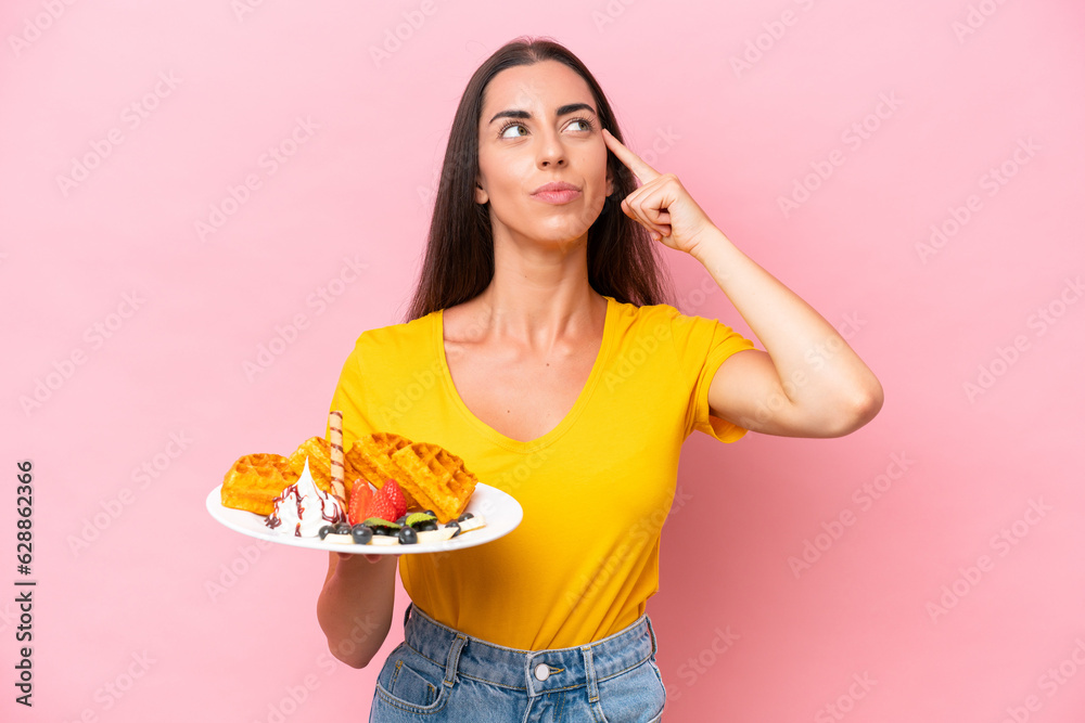 Young caucasian woman holding waffles isolated on pink background having doubts and thinking Young caucasian woman holding waffles isolated on pink background having doubts and thinking