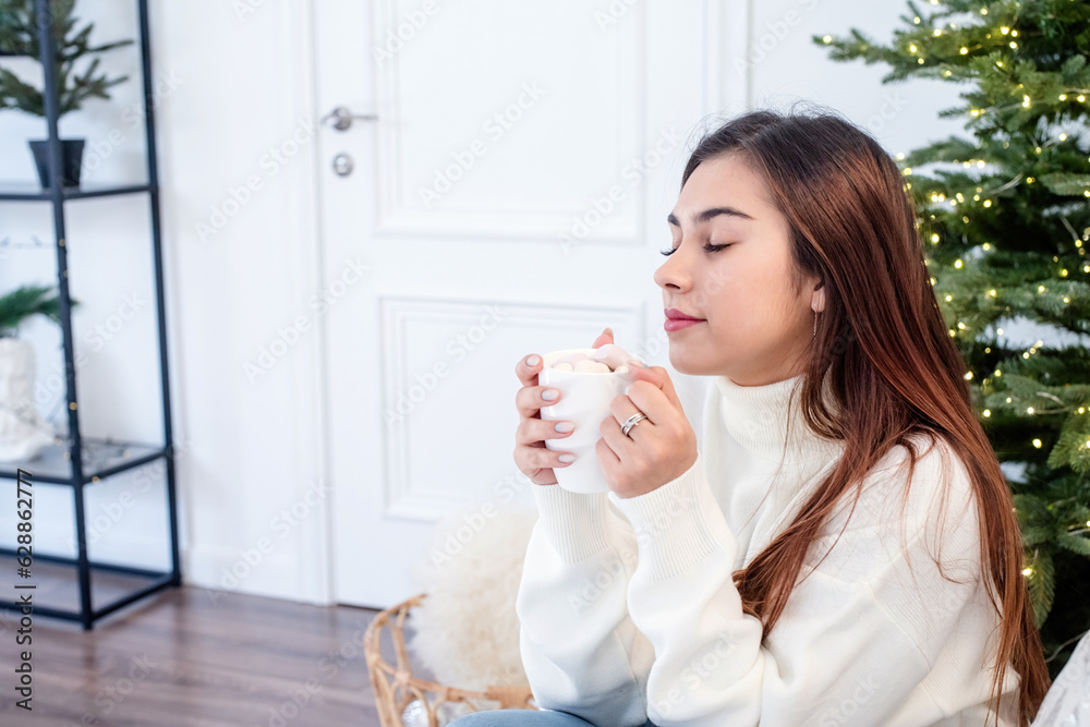 Woman in warm white winter sweater sitting at windowsill at home at christmas eve holding cup with marshmallows Woman in warm white winter sweater sitting at windowsill at home at christmas eve holding cup with marshmallows