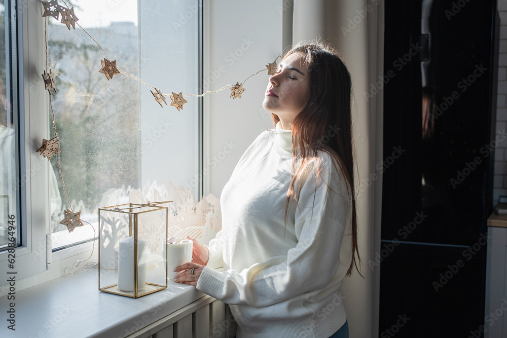 Woman in warm white winter sweater sitting at windowsill at home at christmas eve holding cup with marshmallows