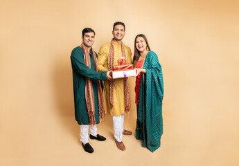 Happy young indian family brother sister wearing traditional cloths holding red gift boxes standing isolated on beige background. Celebrating diwali festival.