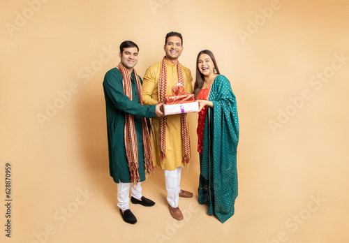 Photographie Happy young indian family brother sister wearing traditional cloths holding red gift boxes standing isolated on beige background