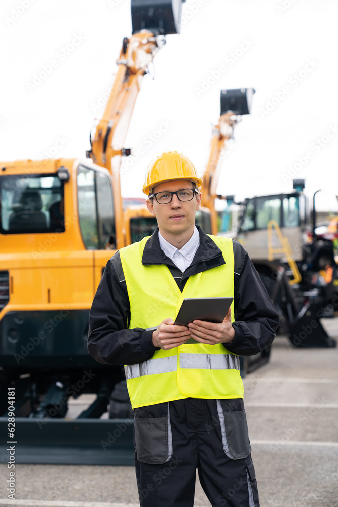 Engineer in a helmet with a digital tablet stands next to construction excavators. Engineer in a helmet with a digital tablet stands next to construction excavators.