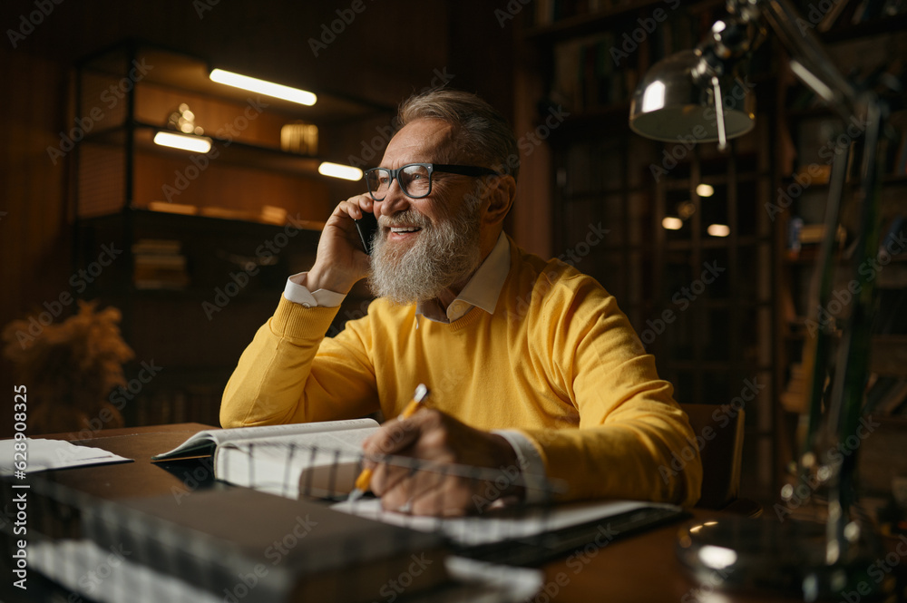 Happy elderly man talking phone while working at home office in evening