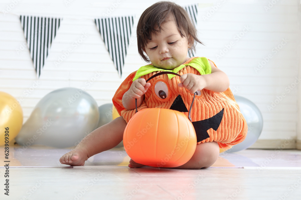 Fototapeta premium Cute baby girl kid dressing up in orange fancy Halloween pumpkin costume, cheerful little cute child holding orange pumpkin to play trick or treat at party. Happy Halloween celebration.