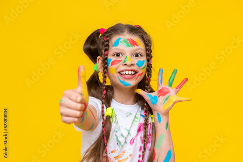 A little girl stained with multicolored paints. A happy, beautiful young girl painted with artistic paints. The child gives a thumbs up and smiles. Yellow isolated background.