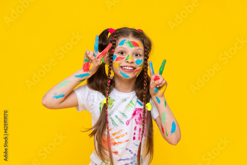 A happy little girl painted with colorful paints. A child with a face smeared with paints, is fond of drawing and shows bunnies from his fingers. Yellow isolated background.