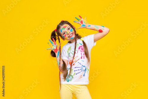 A happy little girl painted with colorful paints. Children's artistic creativity. Painted palms of the hands. A child on a yellow isolated background.