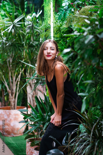 Girl looks straight at camera and smiles. Young woman wearing black dress on thin straps stands among green plants inside arrangement. Young beautiful woman model poses among green tropical plants.