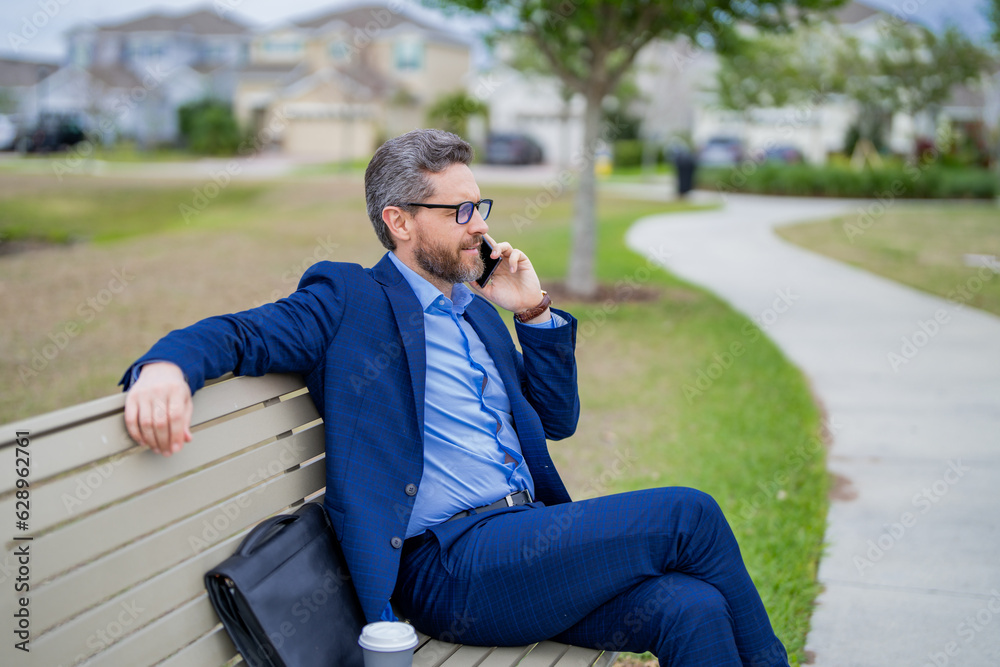 Business man talk on phone sitting on a bench in park. Man in suit call phone outside. Handsome business man using phone sitting on park bench. Business man in suit talk on phone in park. Business man talk on phone sitting on a bench in park. Man in suit call phone outside. Handsome business man using phone sitting on park bench. Business man in suit talk on phone in park.