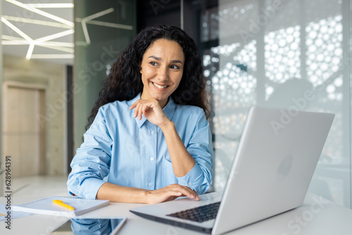 Young beautiful thinking woman smiling and looking out the window, businesswoman smiling at workplace satisfied with achievement results, Latin American woman using laptop at work.