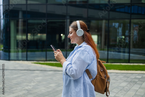 Young happy woman wearing suit using wireless earphones holding smartphone talking on mobile phone having chat on cellphone walking on urban city street. High quality photo