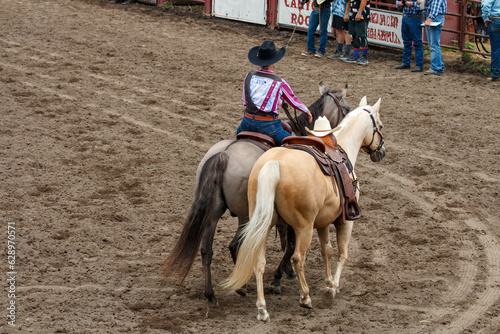 Two horses with one rider is honoring a fallen cowboy. The riderless horse has a white hat on it saddle horn. One horse is white and the other grey. They are in an arena at a rodeo. © Timothy