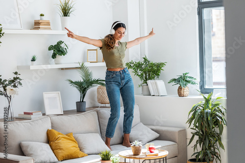 Canvastavla Motivated happy woman singing and dancing on couch while listening music with headphones at home