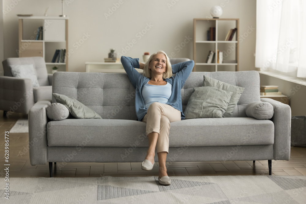 Happy elderly woman relaxing on grey soft couch at cozy home, breathing fresh air, looking at window away, thinking, daydreaming, smiling, laughing, enjoying comfort, leisure in modern interior Happy elderly woman relaxing on grey soft couch at cozy home, breathing fresh air, looking at window away, thinking, daydreaming, smiling, laughing, enjoying comfort, leisure in modern interior