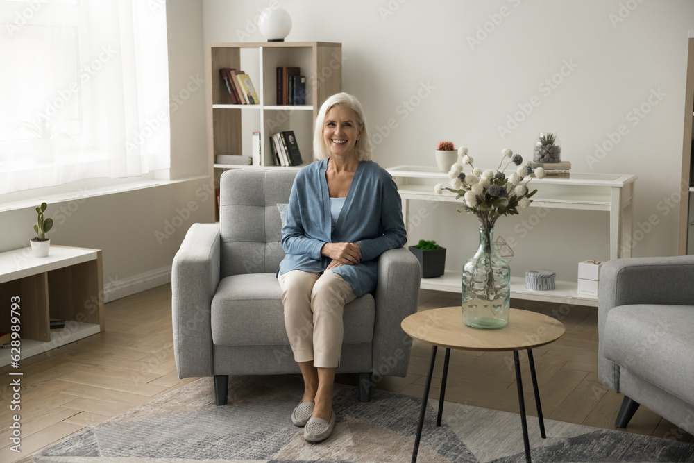 Cheerful pretty blonde senior woman in casual sitting in cozy armchair, looking at camera, smiling, enjoying leisure, posing in modern contemporary home interior. Full length portrait Cheerful pretty blonde senior woman in casual sitting in cozy armchair, looking at camera, smiling, enjoying leisure, posing in modern contemporary home interior. Full length portrait