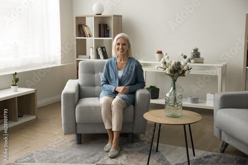 Cheerful pretty blonde senior woman in casual sitting in cozy armchair, looking at camera, smiling, enjoying leisure, posing in modern contemporary home interior. Full length portrait