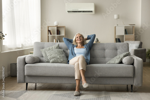 Cheerful relaxed retired old woman resting on grey soft couch under domestic conditioner in stylish cozy apartment interior, breathing cool fresh air, cold comfortable temperature in living room