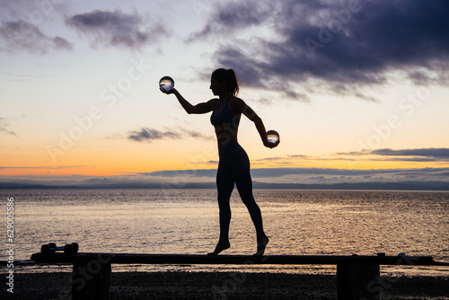 Silhouette of young woman doing workout on a bench with crystal ball(s), in front of ocean at sunrise or sunset