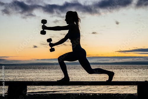 Silhouette of young woman doing workout with weights on a bench in front of ocean sunrise or sunset