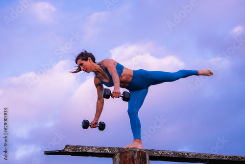 Young woman in blue workout togs, doing workout with weights on a bench in front of ocean sunrise or sunset