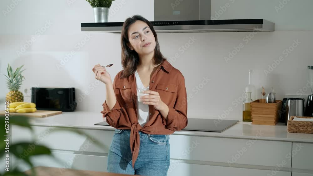 Video of beautiful happy woman eating a yoghurt while looking forwards in the kitchen at home