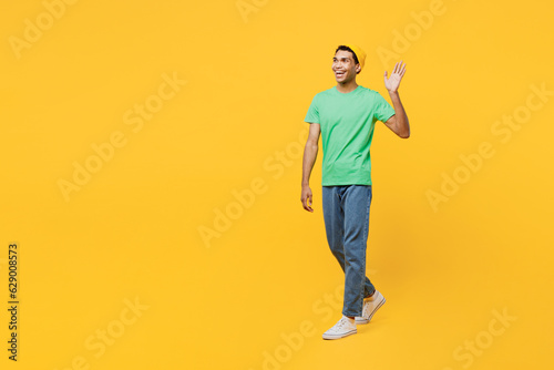Full body smiling fun happy cheerful young man of African American ethnicity he wearing casual clothes green t-shirt hat walking going waving hand isolated on plain yellow background studio portrait.