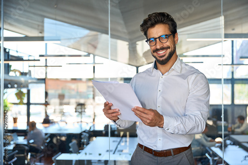 Happy young Latin business man checking financial documents in office. Smiling male professional account manager executive lawyer holding corporate tax bill papers standing at work. Portrait photo