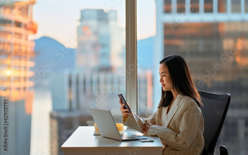 Fotografie Young busy Asian business woman leader holding cellphone device using mobile phone, looking at smartphone checking financial market app working in modern corporate office with big window