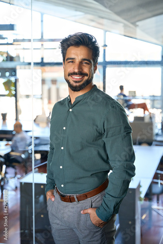 Young Latin business man looking at camera in office, vertical portrait. Smiling happy businessman, male executive manager, professional employee or worker looking at camera standing at work.