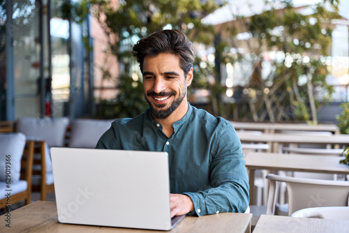 Happy young Latin business man using laptop sitting outdoors. Smiling Hispanic guy student or professional looking at computer sitting in city cafe elearning or hybrid working, searching job online.
