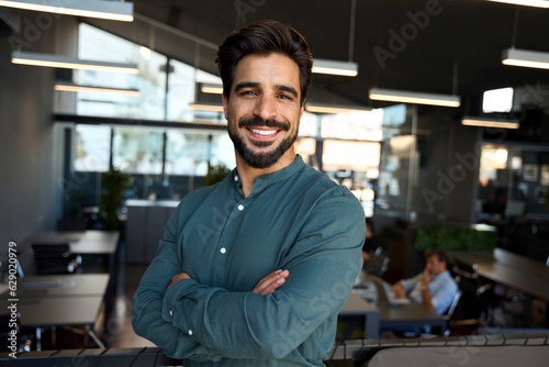 Happy young Latin professional business man looking at camera at work, portrait. Confident smiling bearded businessman, male entrepreneur, company worker or director standing in coworking office.