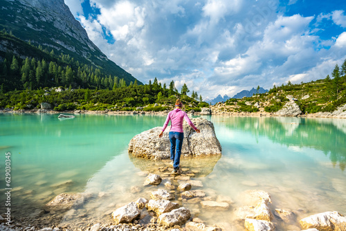 Athletic young woman hops over stone path to large rock at beautiful turquoise Sorapis lake in the afternoon. Lake Sorapis, Dolomites, Belluno, Italy, Europe.