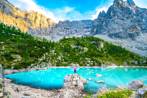 Athletic woman enjoys beautiful view from huge boulder on turquoise Sorapis lake with dito di dio in the evening. Lake Sorapis, Dolomites, Belluno, Italy, Europe.