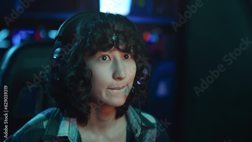 Close-up shot of face of young Asian female gamer with dark curly hair, in headset playing video game in cyberclub and talking into microphone to coordinate with team mates photo