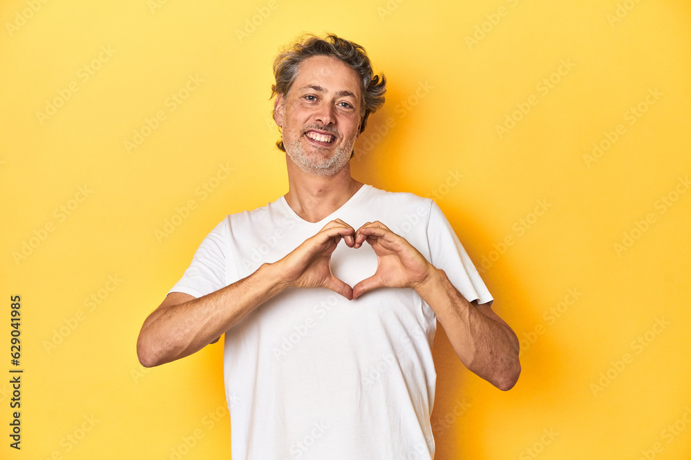 Middle-aged man posing on a yellow backdrop smiling and showing a heart shape with hands. Middle-aged man posing on a yellow backdrop smiling and showing a heart shape with hands.