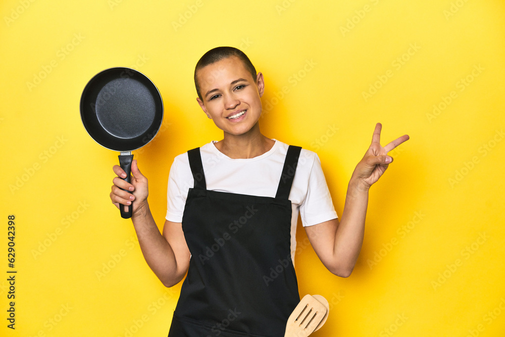 Cook woman holding a pan, yellow studio background joyful and carefree showing a peace symbol with fingers. Cook woman holding a pan, yellow studio background joyful and carefree showing a peace symbol with fingers.